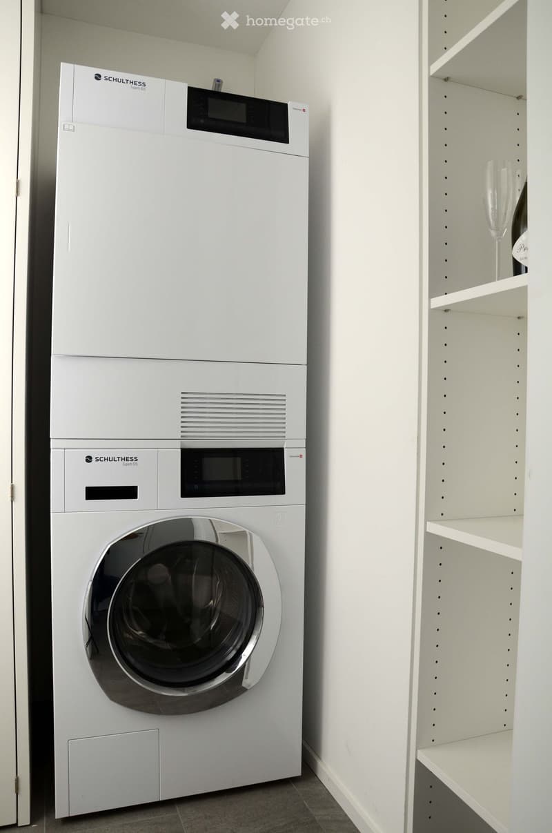 A modern bathroom with a white washing machine and a built-in cupboard with shelves. The washing machine has a sleek, contemporary design with a digital control panel.