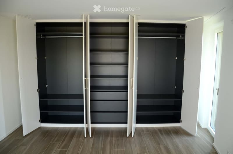 Empty room with wooden flooring and white walls, featuring three large open wardrobes with dark interior shelves and white exterior doors.