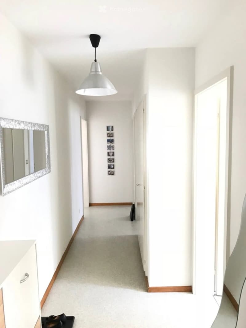 A clean, white-walled hallway with a pendant light fixture, a small cabinet, and a mirror on the left wall. The floor is carpeted, and there are several framed pictures on the wall ahead.