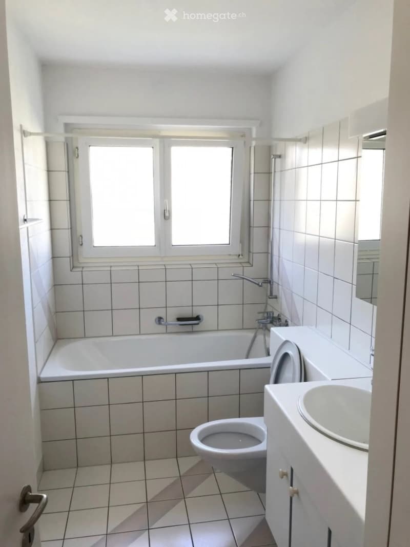 A small bathroom with white tiled walls and floor, featuring a bathtub, toilet, sink, and a window above the bathtub.