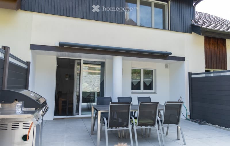 Outdoor patio area with a tiled floor, a dining table with six chairs, and a grill on the left side. The patio is adjacent to a house with large windows and a sliding glass door leading inside.