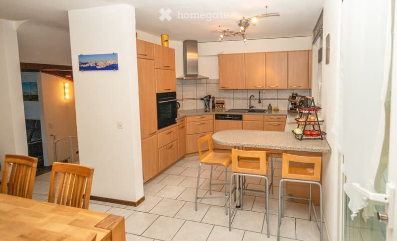 Modern kitchen with light wooden cabinets, stainless steel appliances, granite countertop, and bar stools around a central island.