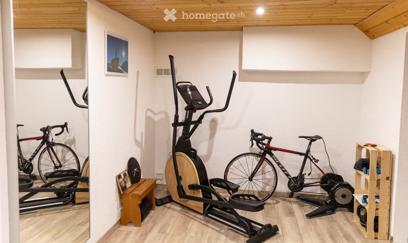 A well-equipped home gym with an elliptical machine, exercise bike, and a wooden bench, featuring light wooden flooring and white walls.