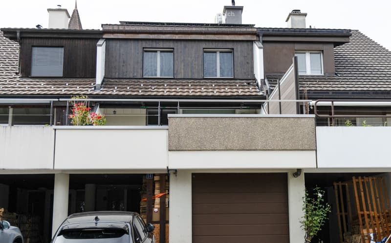 Multi-story residential house with a tiled roof, wooden facade, and balcony with glass railing. Features include a garage, chimney, and small garden area.
