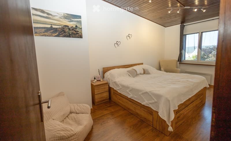 Cozy bedroom with wooden bed, nightstand, armchair, and sofa, featuring a scenic landscape painting on the wall and a view through the windows.