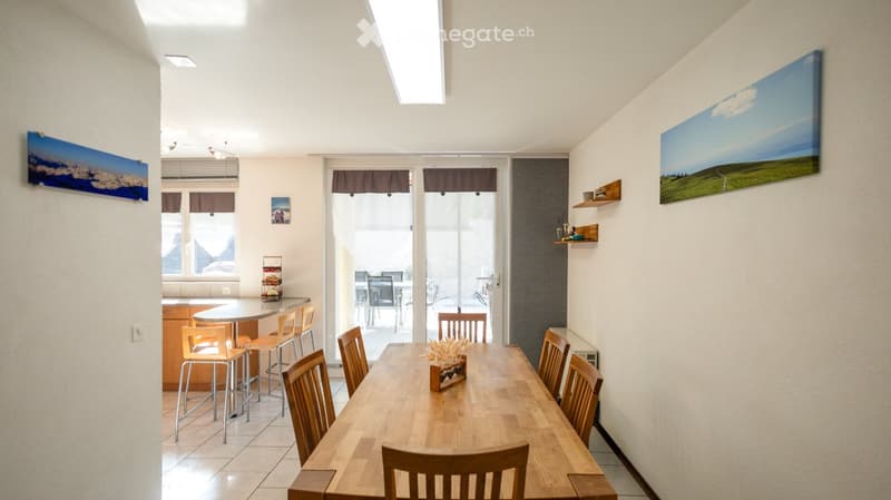Bright dining area with a long wooden dining table surrounded by chairs, a small round table with stools, and a view to a balcony through large glass doors.