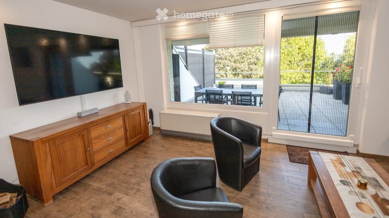 Modern living room with hardwood flooring, large windows, a wooden cabinet, a television, and two black leather chairs.
