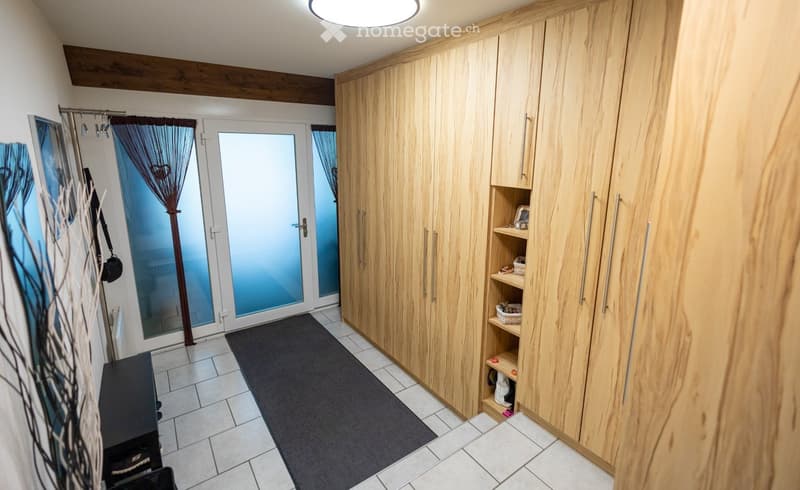 A clean, modern hallway with wooden built-in wardrobes, a dark runner rug, and a glass door leading outside. The ceiling features exposed wooden beams and a round light fixture.