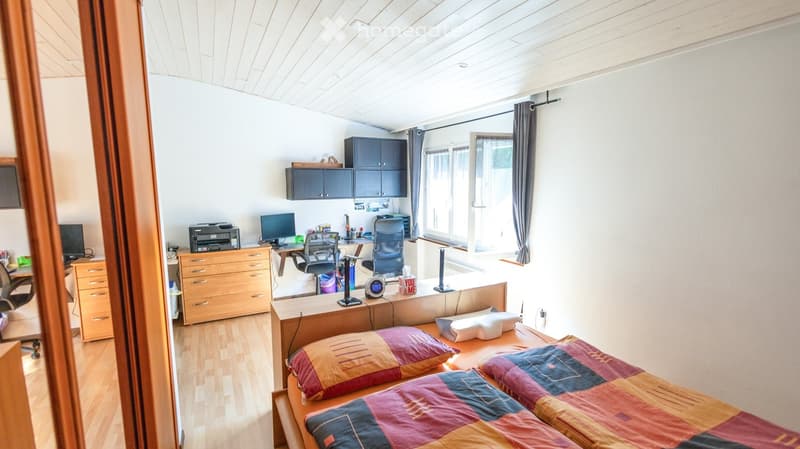 A furnished bedroom with a bed, desk, chair, and cabinet. The room features wooden flooring and large windows with curtains.