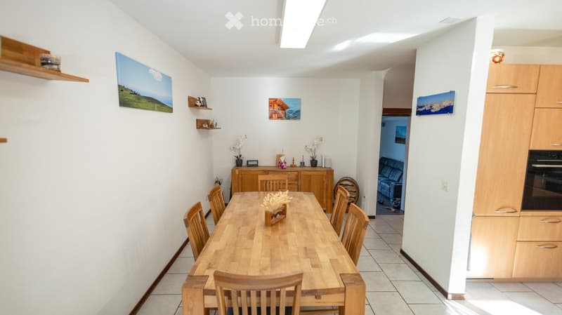 Bright dining area with a wooden dining table surrounded by chairs, a wooden cabinet, and decorative vases on the table. The room features white walls, tiled flooring, and artwork displayed on the walls.