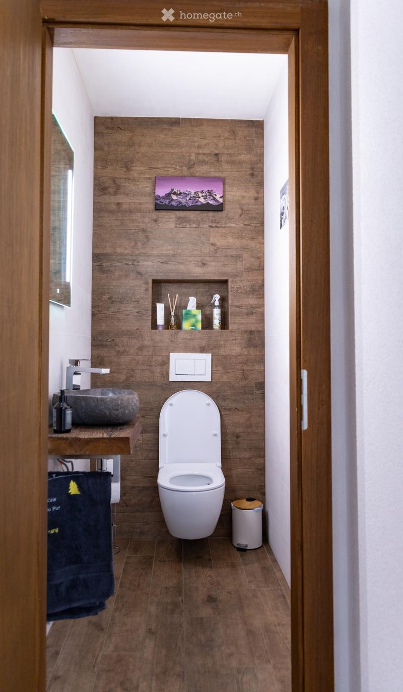 Modern bathroom with wooden walls and floor, featuring a toilet, sink, and a small shelf with toiletries.