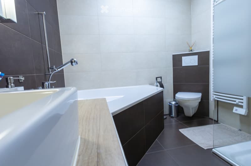 Modern bathroom with a bathtub, sink, toilet, and a glass shower enclosure. The walls are tiled in a combination of dark and light tiles, and the floor is dark tiled.