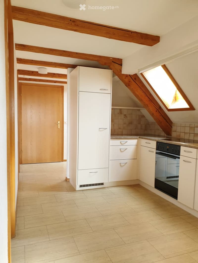 Modern kitchen with white cabinets, a refrigerator, an oven, and exposed wooden beams on the ceiling. The space features light-colored tiled flooring and a skylight window providing natural light.