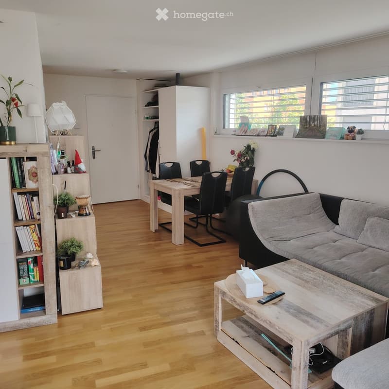 A modern, well-lit living room with a grey sofa, wooden coffee table, dining table with chairs, bookshelf, and decorative items.