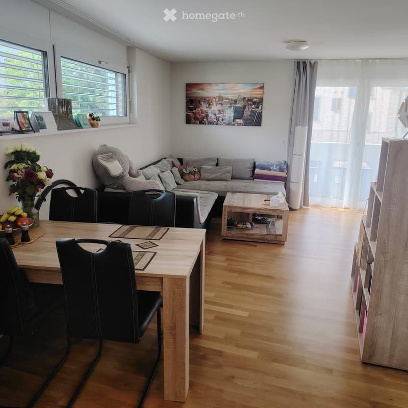 A modern, well-lit living room with a grey sofa, coffee table, dining table with chairs, and a wooden shelf. The room features large windows with sheer curtains, a cityscape painting, and a vase of flowers on the windowsill.