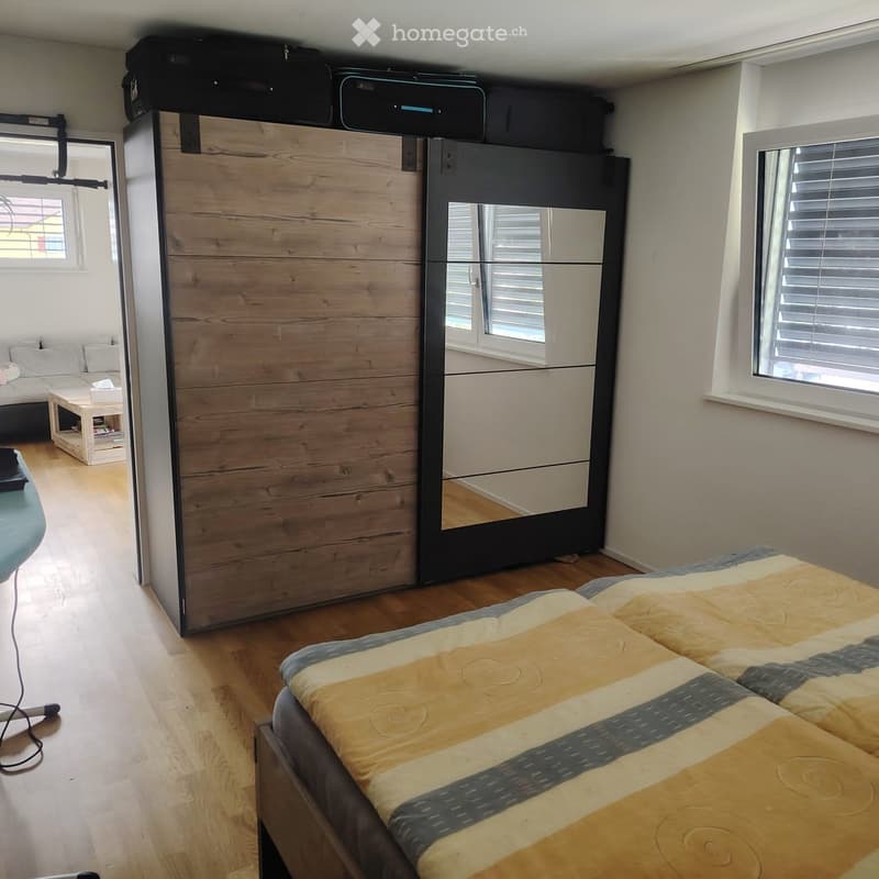 Bright bedroom with wooden flooring, a large bed with a yellow and blue patterned duvet, a modern wardrobe with mirrored doors, a small sofa, and a coffee table.