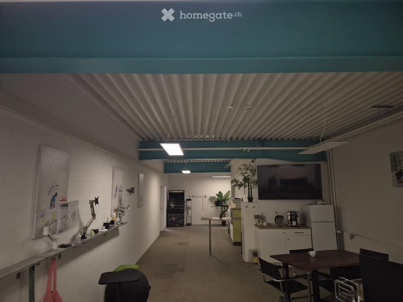 Furnished Room. A modern, open-plan office or lounge area with a mix of furniture including a table, chairs, a refrigerator, a large screen, and a potted plant. The space features exposed ductwork and a mix of white brick and painted walls.