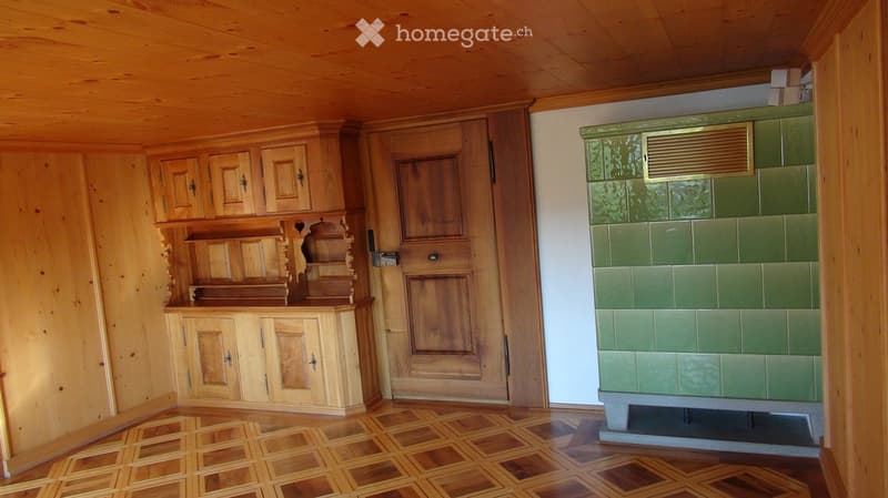 A cozy room with wooden walls, a patterned wooden floor, and a green-tiled fireplace. Wooden cabinets and shelves are mounted on the wall, and a door is visible.