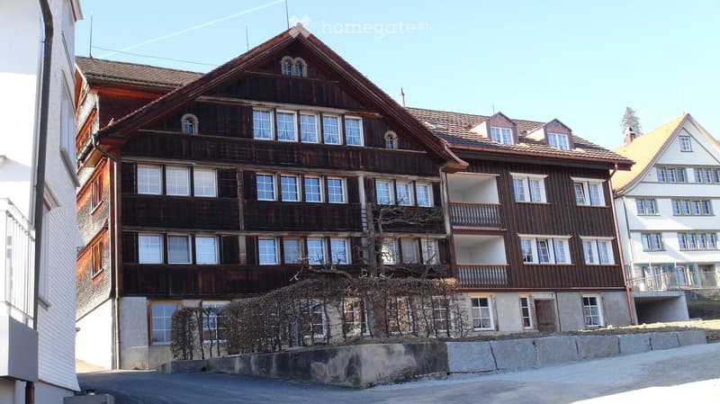 A multi-story wooden building with a steeply pitched roof, multiple windows, and balconies, surrounded by other residential structures.