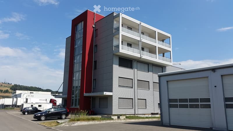 Modern multi-story building with red and grey facade, balconies, and surrounding parking area with several cars.