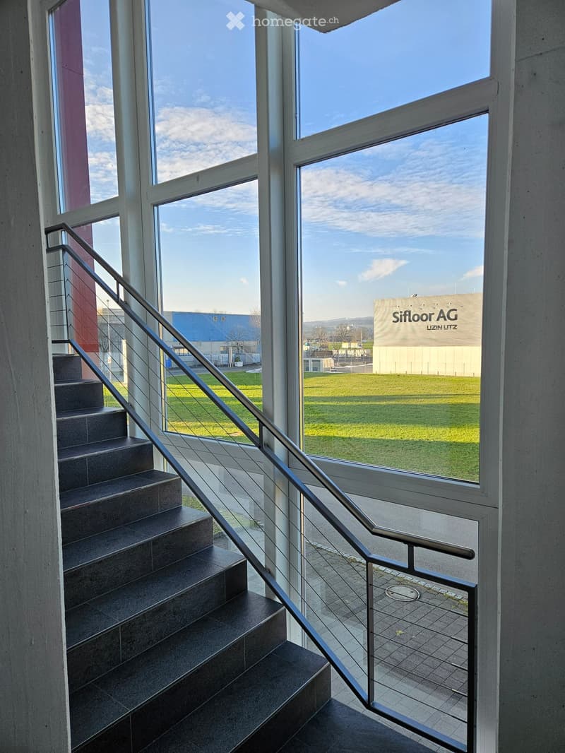 Modern staircase with dark tiled steps and a sleek metal railing, large windows offering a view of a grassy area and industrial buildings.