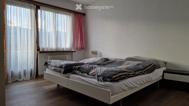Cozy bedroom with a white bed, patterned blankets, wooden flooring, large windows with sheer curtains, and a radiator below the windows.