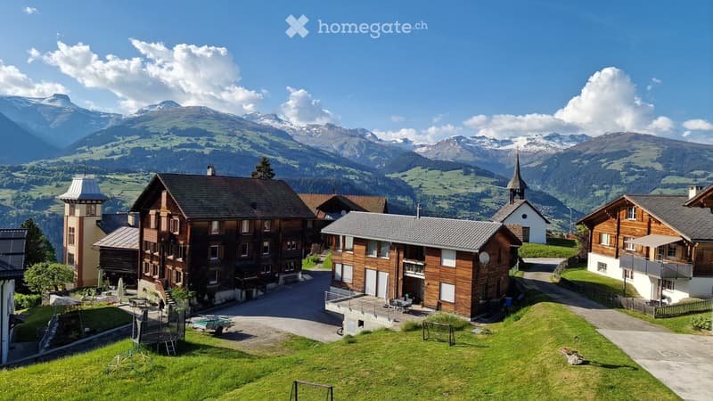 A cluster of traditional wooden houses with pitched roofs and balconies, surrounded by green lawns and a playground, set against a backdrop of snow-capped mountains.