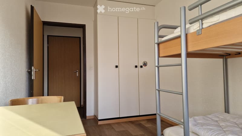 A small, furnished room with a bunk bed, a table, and a chair. The room features light-colored walls, a wooden floor, and built-in white cabinets.