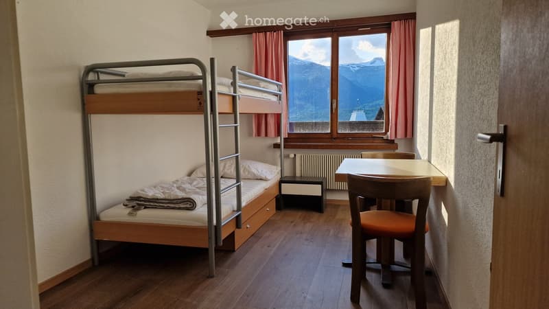 Cozy bedroom with a bunk bed, a small table with a chair, and a nightstand. The room features wooden flooring and large windows with pink curtains, offering a scenic mountain view.