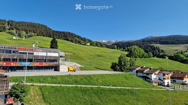 A modern building labeled "Bahn Ober Saxen" sits on a grassy hillside, surrounded by trees and mountains. The structure has a wooden facade with large windows and signage. A few smaller houses are visible in the foreground, and a ski lift runs up the hill