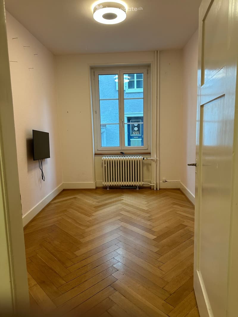 Empty room with herringbone wooden flooring, white walls, a ceiling light fixture, a radiator, and a window with a view of a building outside.