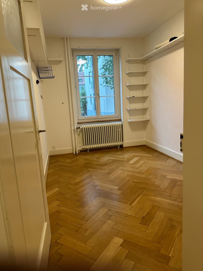 Empty room with wooden herringbone flooring, white walls, built-in shelves, a window with radiator below, and a door on the left side.