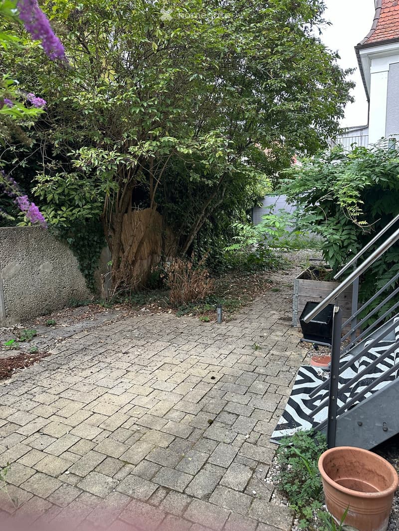 Garden area with paved brick ground, surrounded by greenery and a staircase leading upwards.