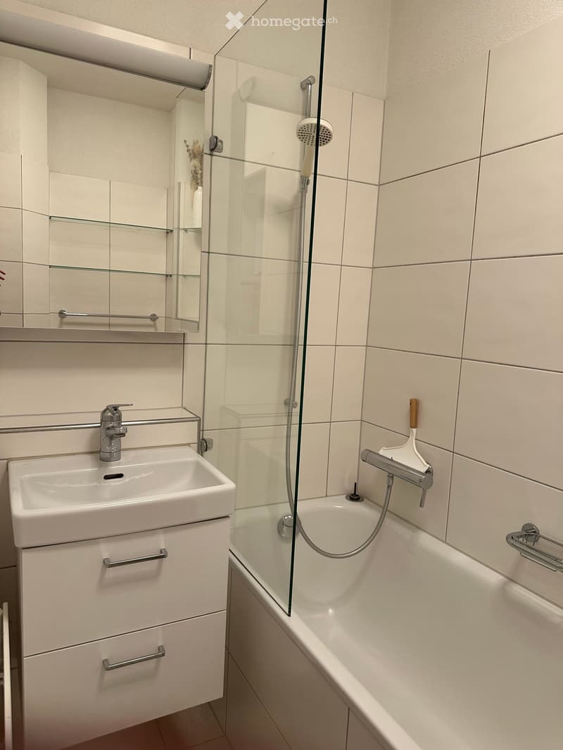 Modern bathroom with white tiles, a sink with a chrome faucet, a bathtub with a glass shower enclosure, and a mirror with glass shelves.