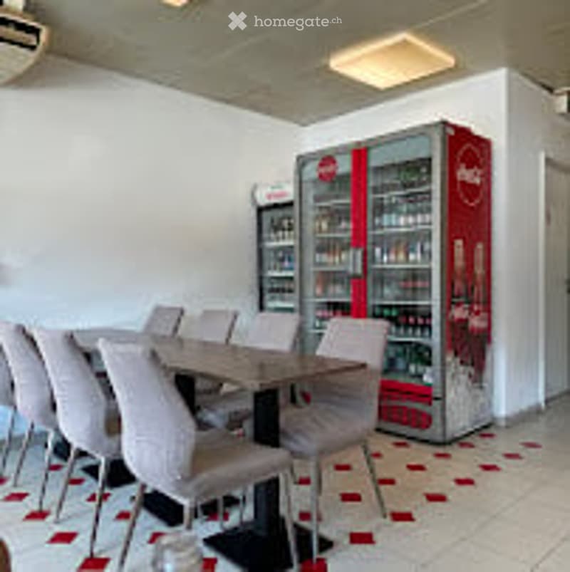 A modern break room with a long table, several chairs, and a refrigerator. The floor has a red and white checkered pattern, and the walls are white.