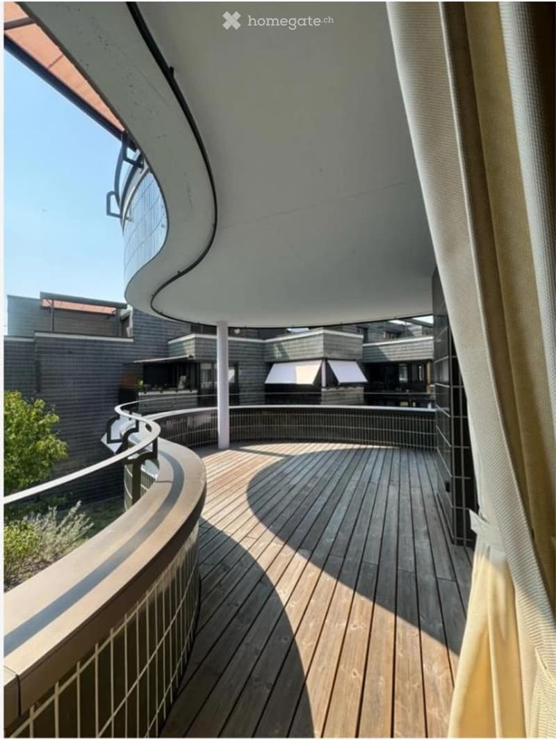 Curved wooden balcony with metal railing, overlooking a modern building complex with grey brick walls and white awnings.
