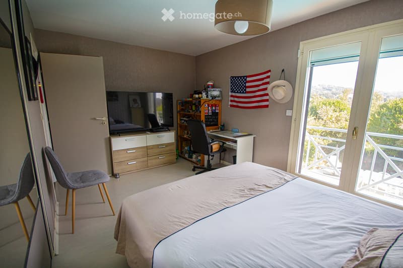 Well-lit bedroom with a bed, desk, chair, television, and a view through large windows.