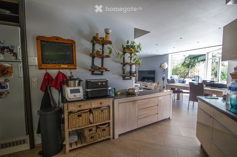 Bright, modern kitchen with light wooden cabinets, stainless steel appliances, and a central island. Features include a refrigerator, microwave, printer, shelves with plants and decorative items, and a dining area with a table and chairs.