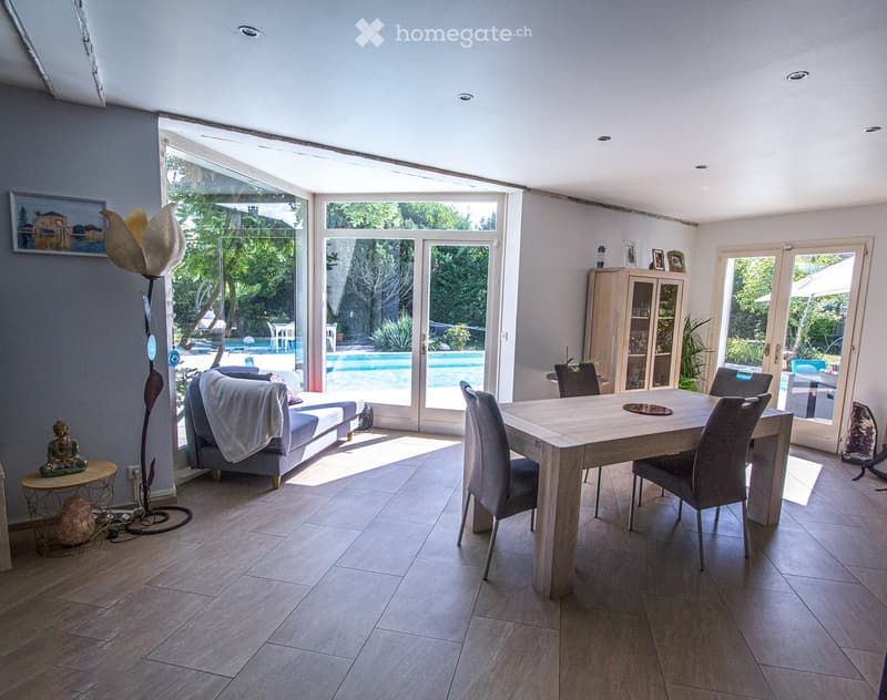 Bright living and dining area with large windows and sliding doors opening to a pool area. Features a sofa, dining table with chairs, a cabinet, a floor lamp, and decorative items.