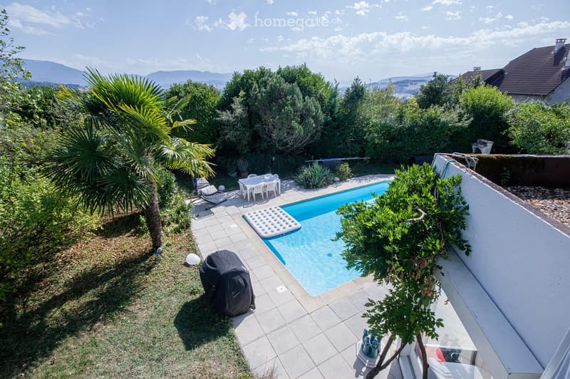 A well-maintained garden with a swimming pool, surrounded by lush greenery, including palm trees and shrubs. There is a covered outdoor seating area with a table and chairs, and a covered barbecue area. The garden features a tiled patio and a grassy area.