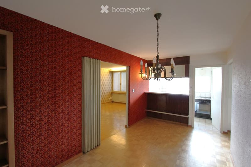 Empty room with wooden flooring, red patterned wallpaper, and a chandelier hanging from the ceiling. The room has an open doorway leading to another room with a window and a radiator.