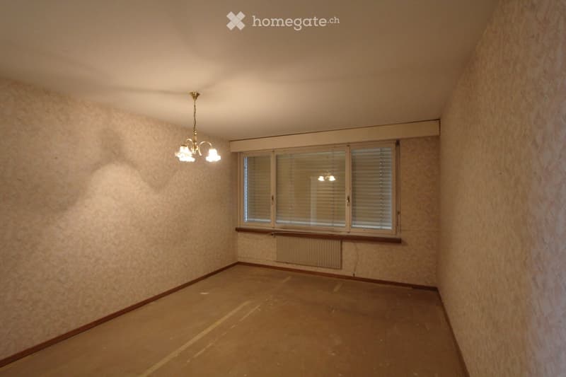 Empty room with beige walls, wooden floor, large window with blinds, radiator, and a chandelier light fixture.
