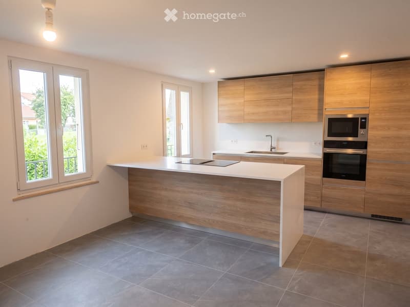 Modern kitchen with light wooden cabinets, white countertops, and stainless steel appliances including a microwave and oven. Features a central island with a built-in induction cooktop and ample natural light from large windows.