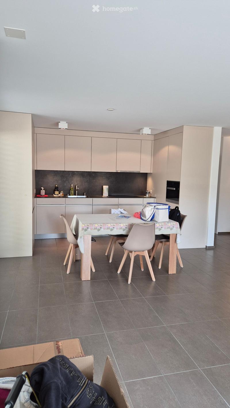 Modern kitchen with light-colored cabinets, a central dining table with chairs, and various items on the counter including bottles, a backpack, and a cup.