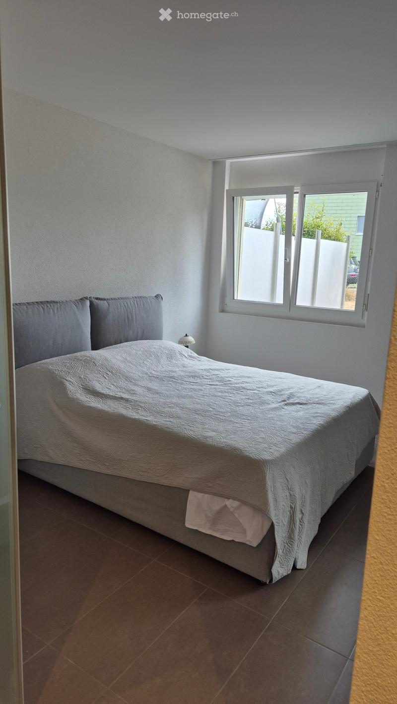 A neatly arranged bedroom with a single bed, grey bedding, and a bedside lamp. The room features tiled flooring and large windows with white frames.