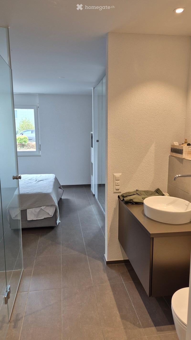 Modern, minimalist room with a bed, a sink, a toilet, and a cabinet. The space features tiled flooring and large windows allowing natural light.