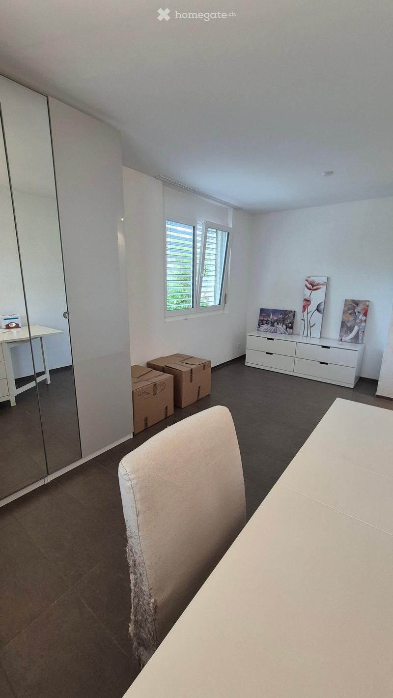 A furnished room with a large window, a white cabinet with drawers, a mirror, a chair, a table, and several cardboard boxes on the floor.