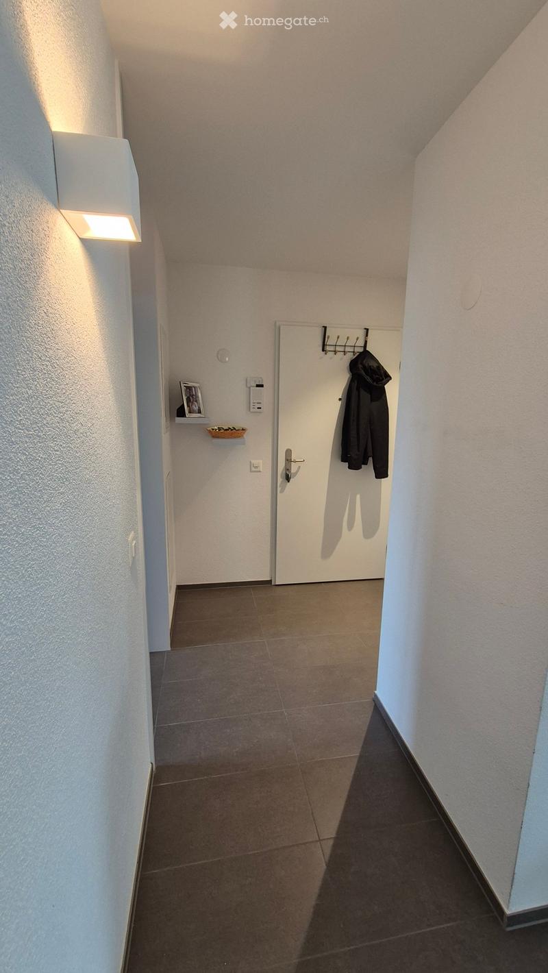 A narrow hallway with tiled flooring, white walls, and a coat rack holding a jacket. A small shelf with a framed photo and a decorative bowl is visible on the left side.