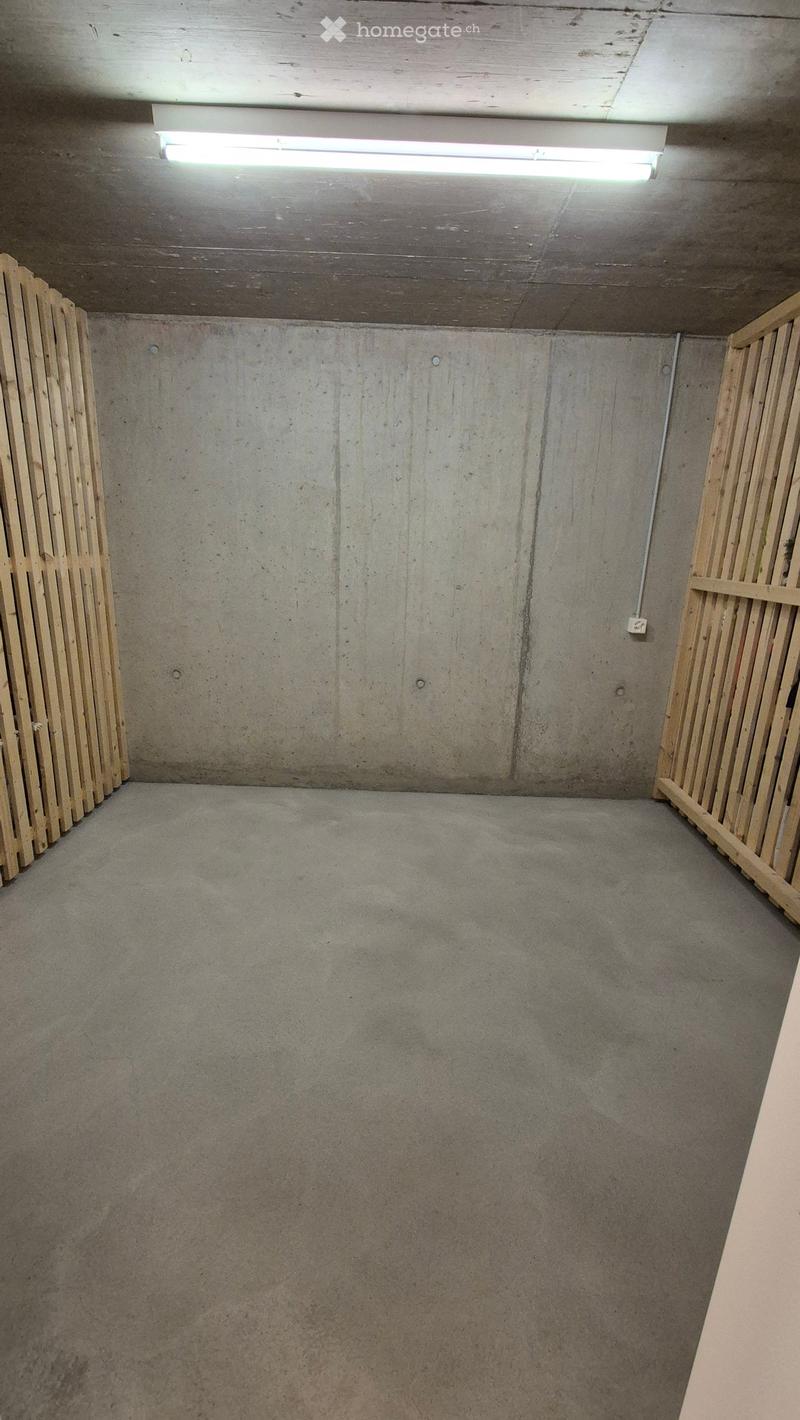 Empty concrete-walled room with a single overhead light fixture and wooden planks stacked against the walls.
