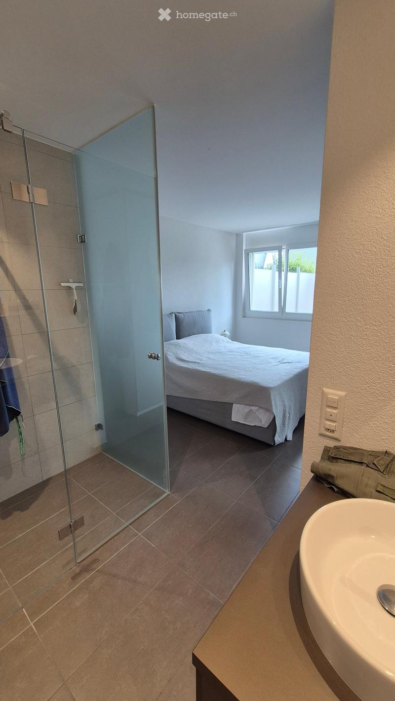Modern bedroom with a large bed, a glass-enclosed shower, and a sink in the corner. The room features tiled flooring and light-colored walls.